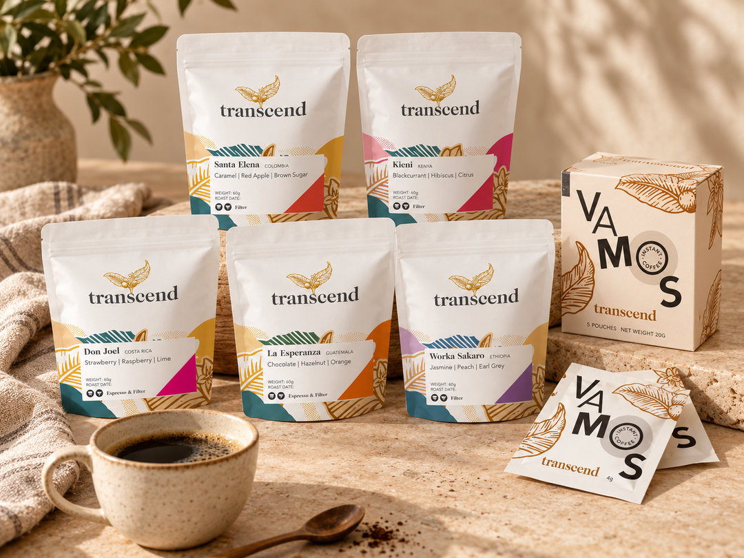 Six Transeend coffee pouches in assorted flavors are arranged on a wooden surface with a matching branded box, a cup of black coffee, a spoon, and two individual tea bags. The background is a warm, softly blurred indoor setting with a plant and neutral-toned wall.