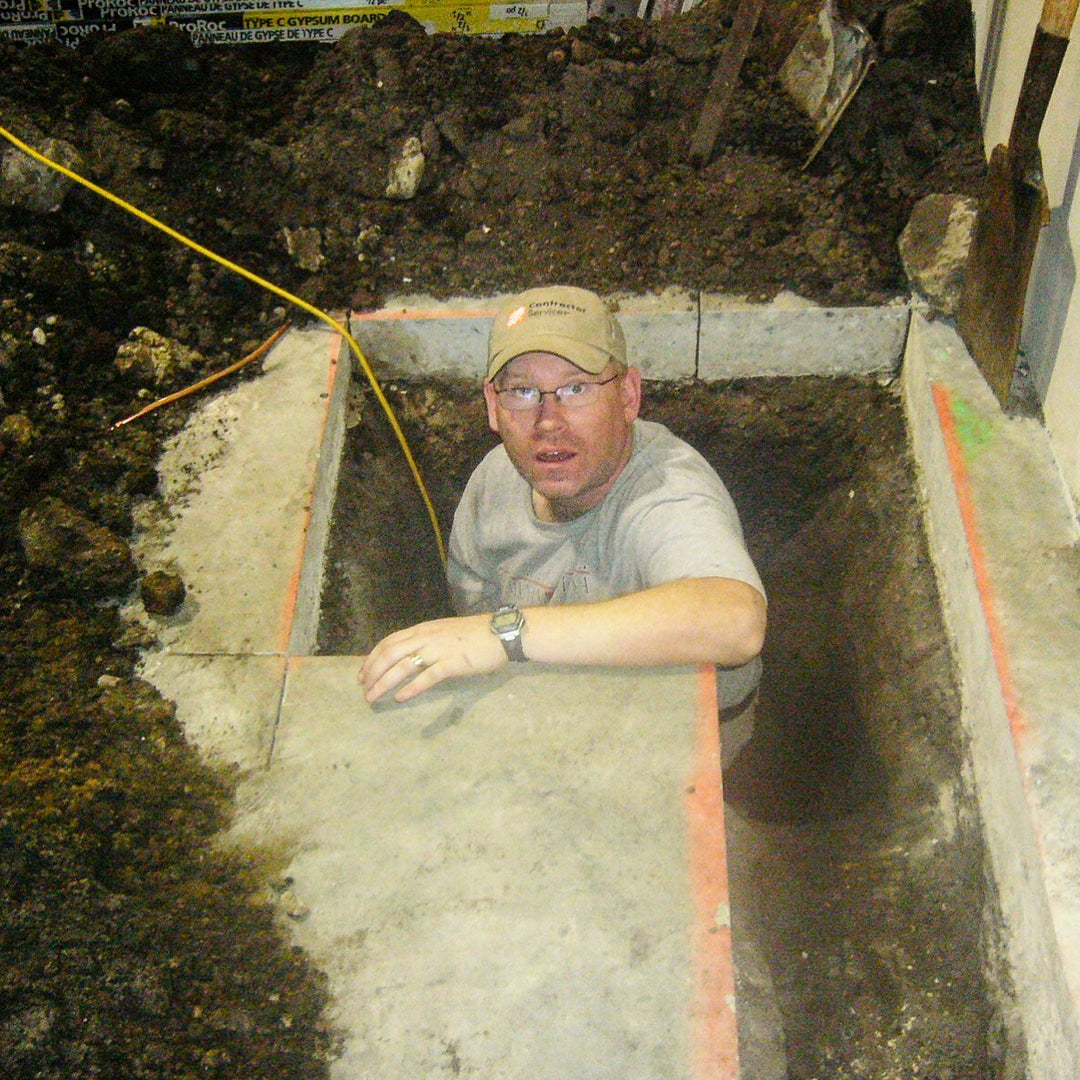 Poul in a cap and glasses sitting inside a dug-out trench with soil and construction materials around.